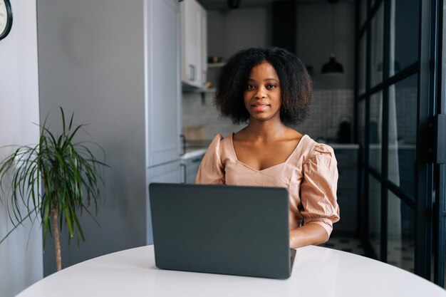 Portret van Afro-Amerikaanse jonge zakenvrouw zittend aan tafel in moderne keuken kamer werken op laptop glimlachend camera kijken