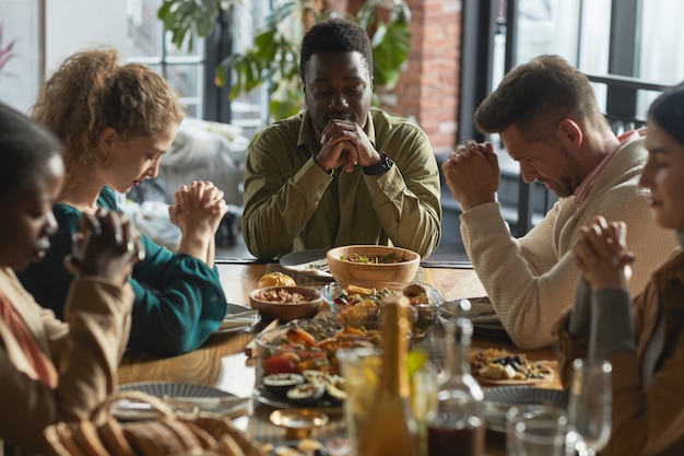Portret van Afro-Amerikaanse man bidden met ogen dicht zittend aan tafel tijdens Thanksgiving-viering met vrienden en familie,