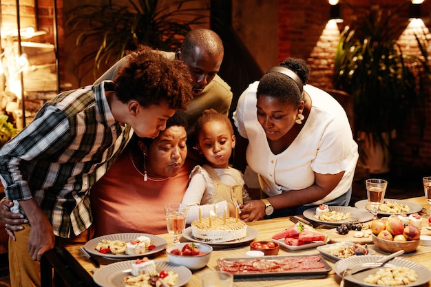 Portret van Afro-Amerikaanse oma die kaarsen blaast terwijl ze verjaardag viert met familie in gezellige ...