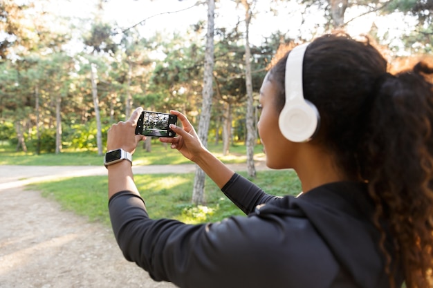 Portret van Afro-Amerikaanse vrouw 20s dragen zwarte trainingspak en koptelefoon, selfie foto nemen op mobiele telefoon tijdens een wandeling door groen park