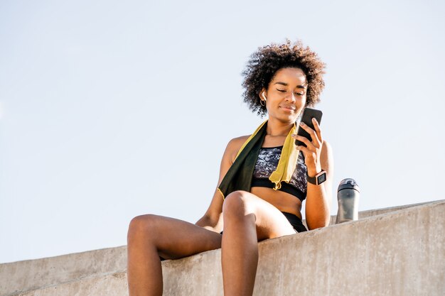 Portret van afro atleet vrouw met behulp van haar mobiele telefoon en ontspannen na het trainen buitenshuis