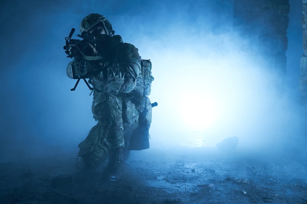 Portret van airsoft-speler in professionele uitrusting met machinegeweer in verlaten verwoeste gebouw. Soldaat met wapens in oorlog in rook en mist