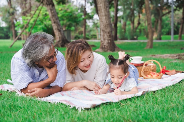 Portret van Aziatische grootouders en kleindochter die op het groene glasgebied openlucht leggen, familie die van picknick samen in het concept van de de zomerdag genieten