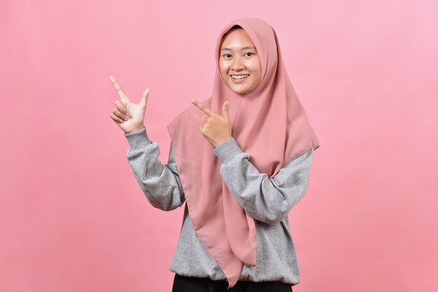 Portret van Aziatische mooie jonge moslimvrouw die beide handen met de vinger aan de bovenzijde wijst met een glimlach, geïsoleerd op roze achtergrond