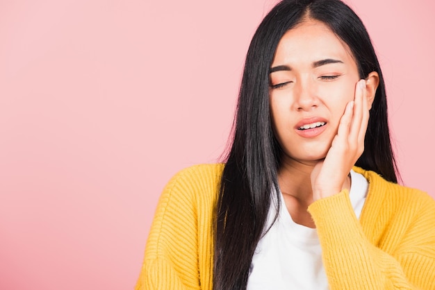 Portret van Aziatische mooie jonge vrouw die lijdt aan kiespijn, vrouwelijke verschrikkelijke sterke tanden pijn probleem hand op kin te drukken, studio opname geïsoleerd op roze achtergrond, tandheelkundig zorgconcept