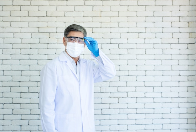 Portret van aziatische oudere arts of onderzoeker dragen laboratoriumjas en gezichtsmasker staande en hand houden eyeglassed met witte bakstenen achtergrond.