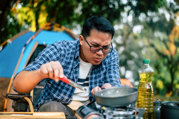 Portret van Aziatische reiziger man bril varkensvlees steak frituren BBQ in braadpan pan of pot op een camping buiten koken reizen camping lifestyle concept