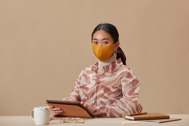 Portret van Aziatische vrouw in beschermend masker voorzijde kijken zittend aan tafel en met behulp van digitale tablet