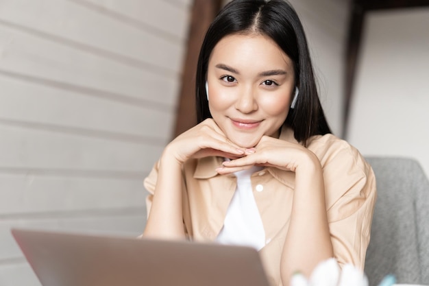 Portret van aziatische vrouw in oortelefoons die aan laptop werkt en glimlacht die gelukkig naar camera kijkt die studeert ...