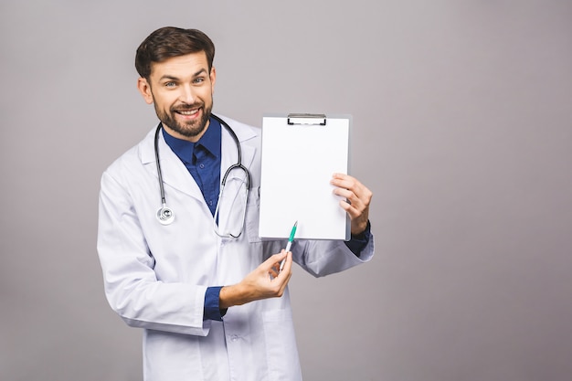 Portret van bebaarde lachende stagiair, die het klembord met leeg papier houdt. dokter draagt witte uniform, staat over geïsoleerde grijze achtergrond, met blanco.