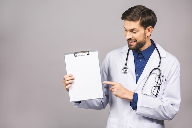 Portret van bebaarde lachende stagiair, die het klembord met leeg papier houdt. Dokter draagt witte uniform, staat over geïsoleerde grijze achtergrond, met blanco.