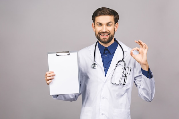 Portret van bebaarde lachende stagiair, die het klembord met leeg papier houdt. Dokter draagt witte uniform, staat over geïsoleerde grijze achtergrond, met blanco.