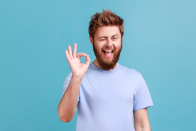 Portret van bebaarde man oke teken succes teken tonen tevreden met goede service camera kijken en knipogen uiten van positieve emoties Indoor studio shot geïsoleerd op blauwe achtergrond