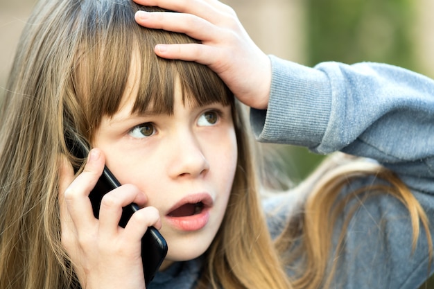 Portret van beklemtoond kindmeisje met lang haar dat op celtelefoon spreekt. Kleine vrouwelijke jongen communiceren via smartphone. Kinderen communicatieconcept.