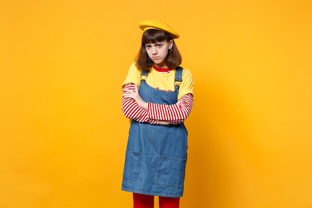 Portret van bezorgd beledigd meisje tiener in Franse baret denim zomerjurk hand in hand gevouwen geïsoleerd op gele achtergrond in studio. Mensen oprechte emoties levensstijl concept. Bespotten kopie ruimte.