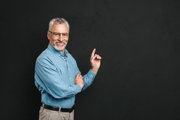 Portret van blanke gepensioneerde man jaren '60 met grijs haar en baard in overhemd glimlachend en wijsvinger opzij wijzen op copyspace, geïsoleerd over zwarte muur