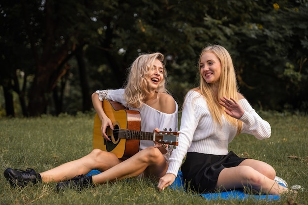 Portret van blanke jonge vrouwen die buiten in het park zitten en gitaar spelen, zingen samen met geluk een lied