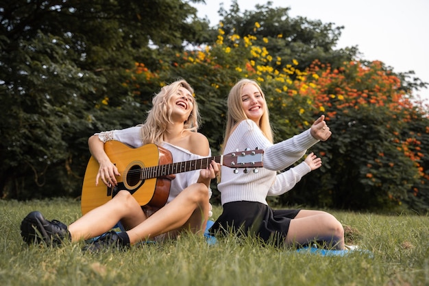 Portret van blanke jonge vrouwen die buiten in het park zitten en gitaar spelen, zingen samen met geluk een lied