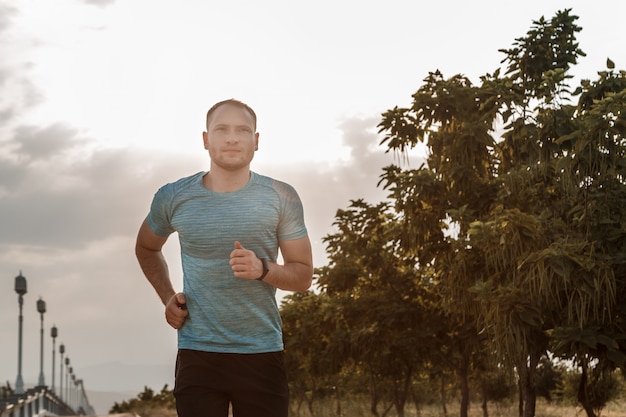 Portret van blanke man in een blauw t-shirt en zwarte korte broek die tijdens zonsondergang op het asfaltspoor traint en loopt