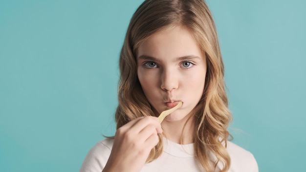 Portret van blond tienermeisje dat haar tanden schoonmaakt op camera geïsoleerd op blauwe achtergrond