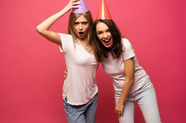 Portret van blonde en brunette jonge vrouwen close-up met verjaardag hoeden plezier geïsoleerd op roze achtergrond.