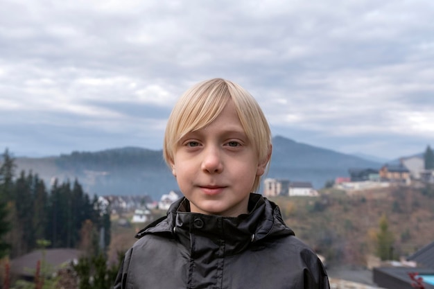Portret van blonde jongen in zwarte jas op de bergen achtergrond Blonde tiener buiten Schooljongen loopt naar buiten
