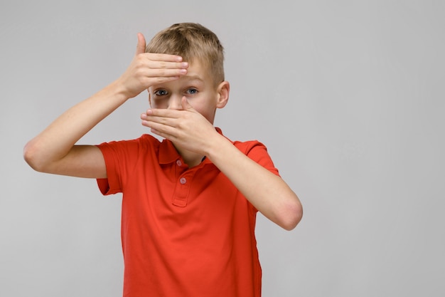 Portret van blonde Kaukasische droevige zieke kleine jongen in oranje t-shirt die zijn voorhoofd en mond met handen sluiten