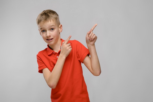 Portret van blonde Kaukasische ernstige kleine jongen in oranje t-shirt tonen op lege ruimte op grijze achtergrond