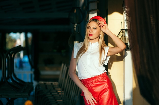 Portret van blonde vrouw in rode baret en lederen rode broek poseren buiten. Rode bril en sjaal.