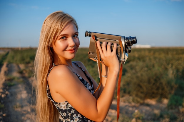 Portret van blondevrouw in bloemendrukkleding met uitstekende videocamera op druivengebied tijdens zonsondergang