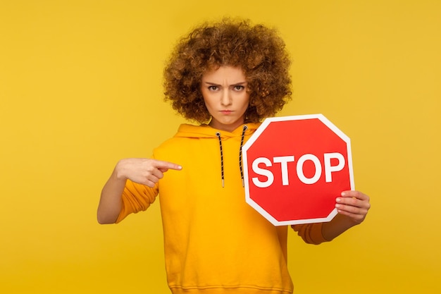 Foto portret van boos krullend meisje wijzend rood stopbord en kijken naar camera met negatieve agressieve uitdrukking weergegeven: verbod verbod symbool indoor studio shot geïsoleerd op gele achtergrond