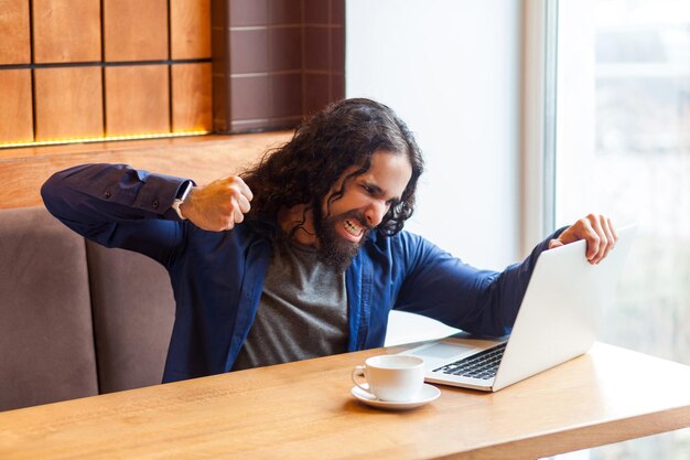 Portret van boze agressieve jonge volwassen man freelancer in casual stijl zittend in café en proberen te crashen laptop scherm opgeheven vuist met gebalde tanden Indoor lifestyle concept