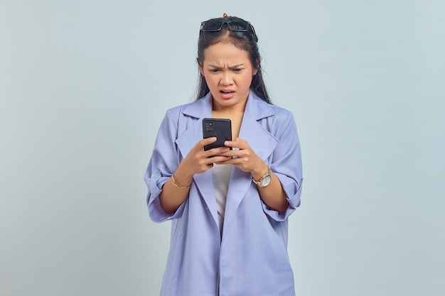 Portret van boze jonge Aziatische vrouw die mobiele telefoon gebruikt die op witte achtergrond wordt geïsoleerd