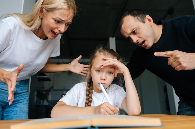 Portret van boze jonge ouders schreeuwen en schelden samen lui dochtertje zittend aan tafel huiswerk triest camera kijken