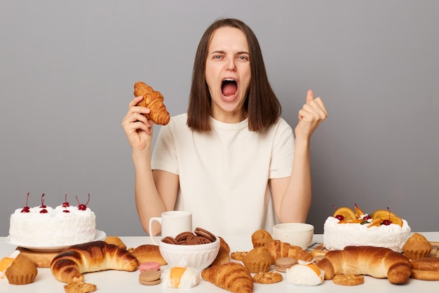 Portret van boze wanhoop vrouw met bruin haar dragen witte T-shirt zittend aan tafel geïsoleerd over grijze achtergrond croissant vasthouden en schreeuwen van woede en gebalde vuist