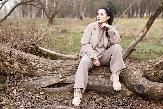Portret van brunette vrouw in beige jas met mooi lang haar zittend op een boom in het park