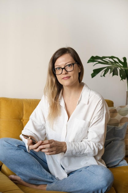 Portret van brunette zakenvrouw freelancer van middelbare leeftijd met behulp van telefoon zittend met benen op geel zo