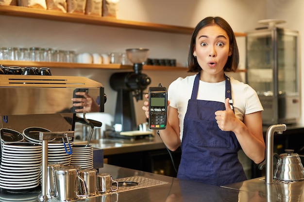 Portret van café barista meisje toont duimen omhoog en kaartlezer betaalautomaat beveelt contactloos aan