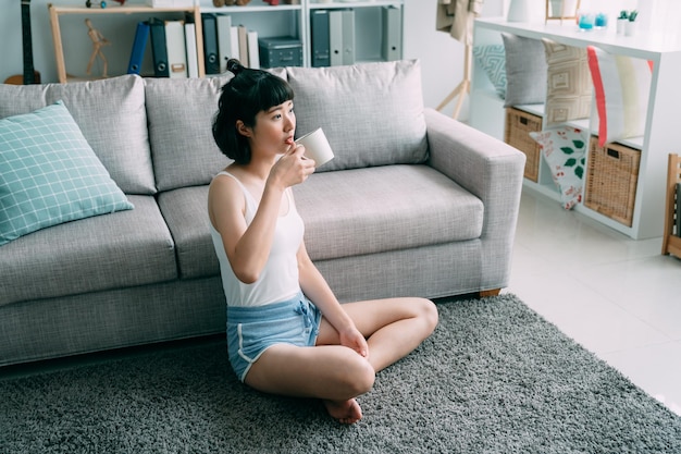 Portret van charmante aziatische chinese vrouw in slaap draagt ontspannen thuis met een kopje thee in de zomer. dame zittend op tapijt in huis woonkamer geniet van koffie in de ochtend kijkt uit raam zonnig