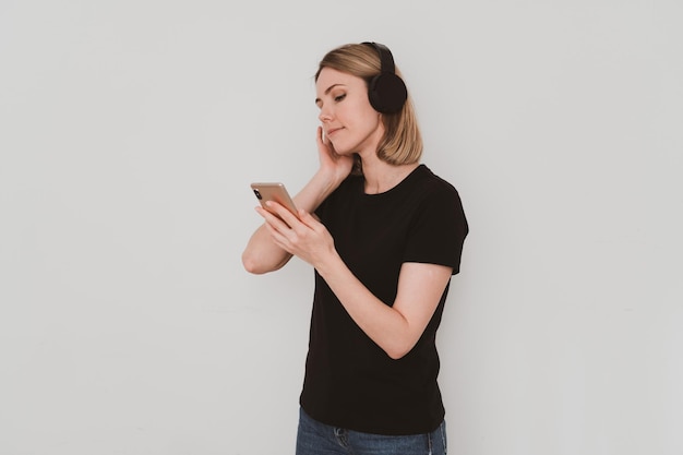 Portret van charmante Europese vrouw in casual kleding met koptelefoon luisteren naar muziek op smartphone geïsoleerd op een witte achtergrond.