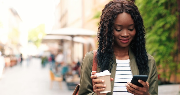 Foto portret van de aantrekkelijke jonge multiraciale vrouw die smartphone praat en buiten koffie drinkt...