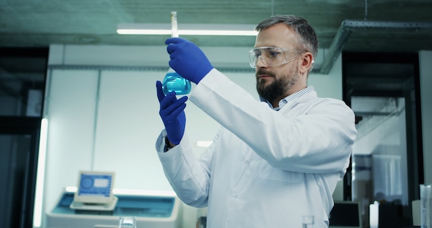 Portret van de blanke man in glazen en witte robe bezig met een analyse van de blauwe vloeistof in de reageerbuis tijdens medisch of farmaceutisch onderzoek in het laboratorium.