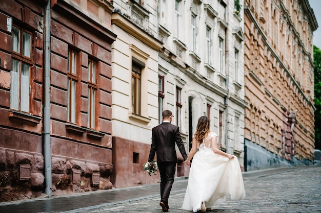 Portret van de bruidegom in trouwpak en de bruid in jurk die terugloopt in de buurt van oud gebouw, oud huis buiten, buiten. Pasgetrouwden lopen door de straten van de stad Lviv. Bruiloft wandelingen.