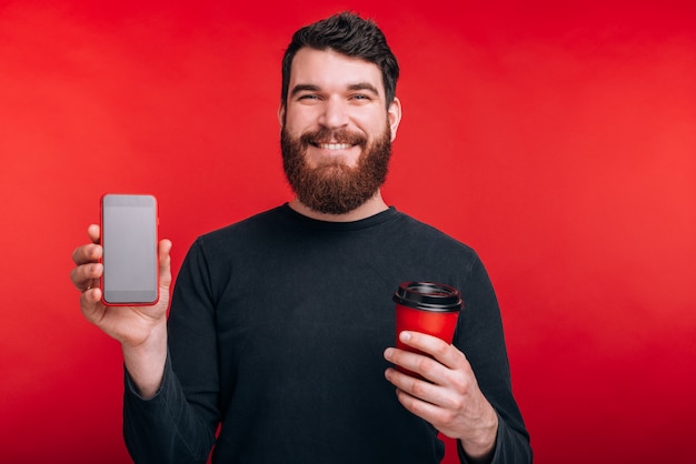 Portret van de glimlachende mens met de kop van de baardholding van koffie om te gaan en het smartphonescherm tonen