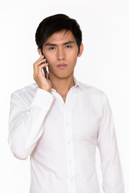 Portret van de jonge knappe Aziatische mens die op mobiele telefoon spreekt die tegen witte muur wordt geïsoleerd