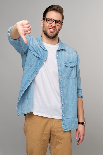 Portret van de jonge knappe man in jeansoverhemd die grote duim omlaag tonen die een hekel hebben aan gebaar dat zich over lichte muur bevindt