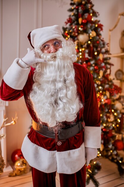 Portret van de kerstman met een bril bij de open haard en de kerstboom met geschenken. Nieuwjaar en vrolijk kerstfeest, gelukkig vakantieconcept