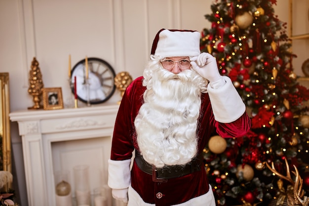 Portret van de kerstman met een bril bij de open haard en de kerstboom met geschenken.