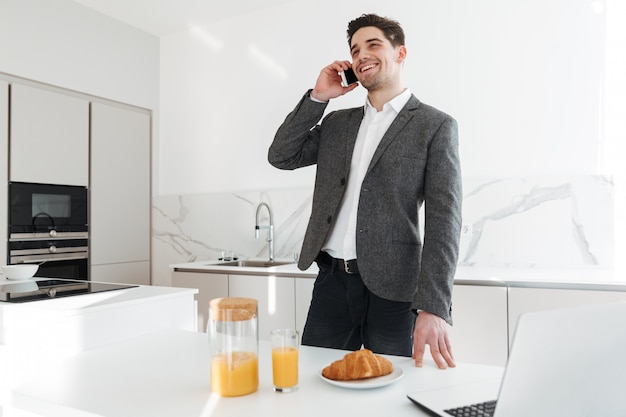 Portret van de succesvolle zakelijke mens die op mobiele telefoon spreken terwijl het hebben van ontbijt en thuis het werken aan laptop