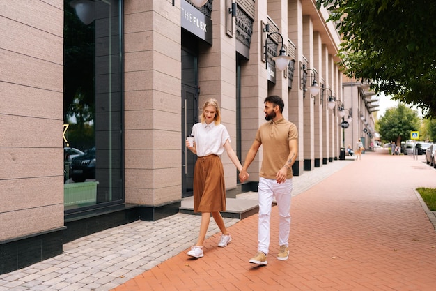 Portret van de volledige lengte van een gelukkig jong verliefd stel dat hand in hand loopt op straat in de stad Stijlvolle bebaarde man en blonde vrouw met kopje met afhaalkoffie